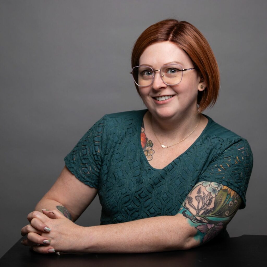 Portrait of Eve Kinney on a gray background. Eve is seated, smiling, with arms in front of her and hands clasped. Eve is a white woman with red hair, glasses, tattoos, and wears a teal shirt.